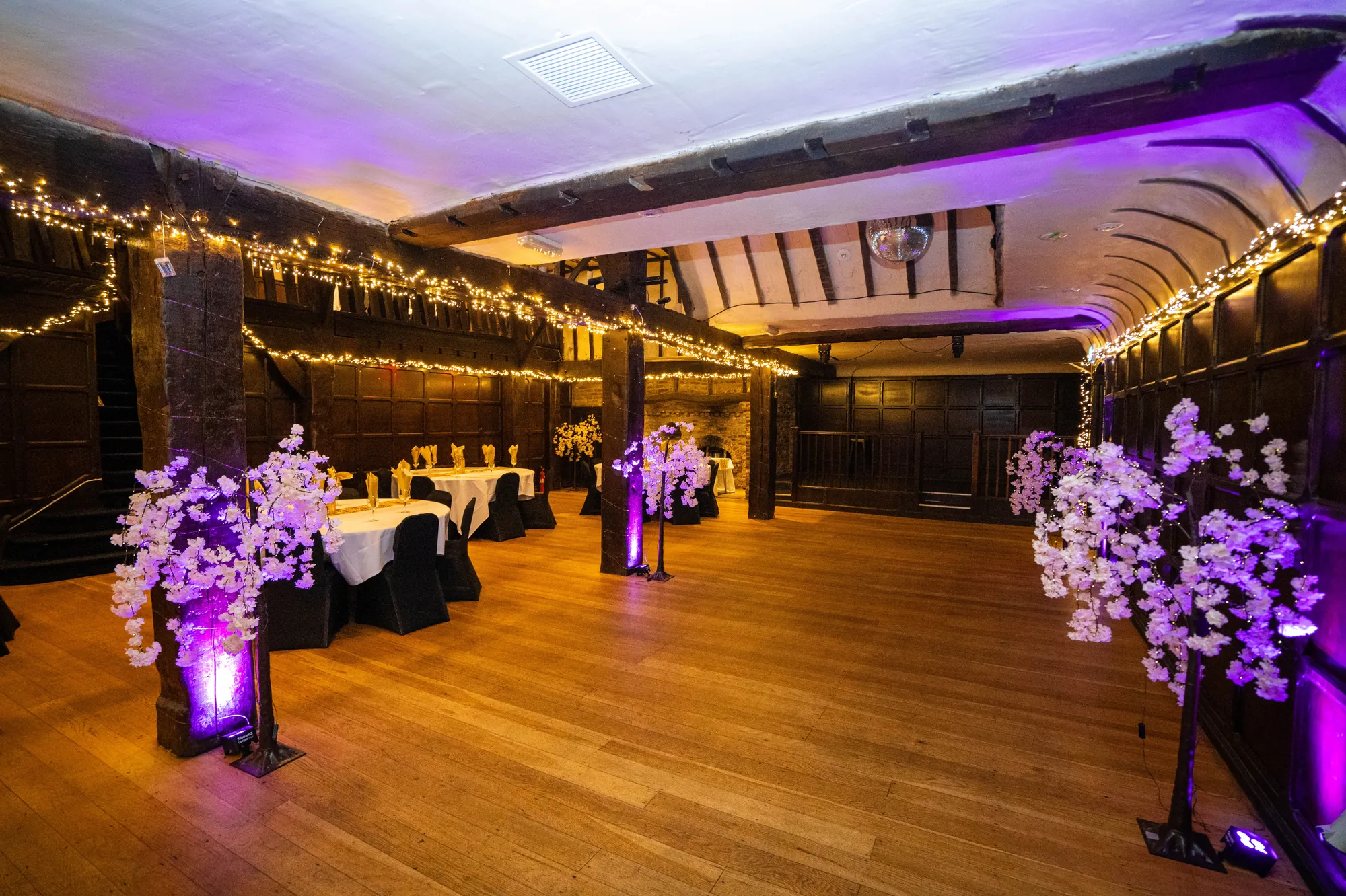 The Baronial Hall set for a wedding, with round tables, purple-lit blossom trees and fairy lights along the beams